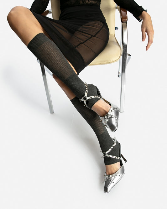 Silver spiked leather heels styled on a seated model with a black skirt and grey knee-high socks